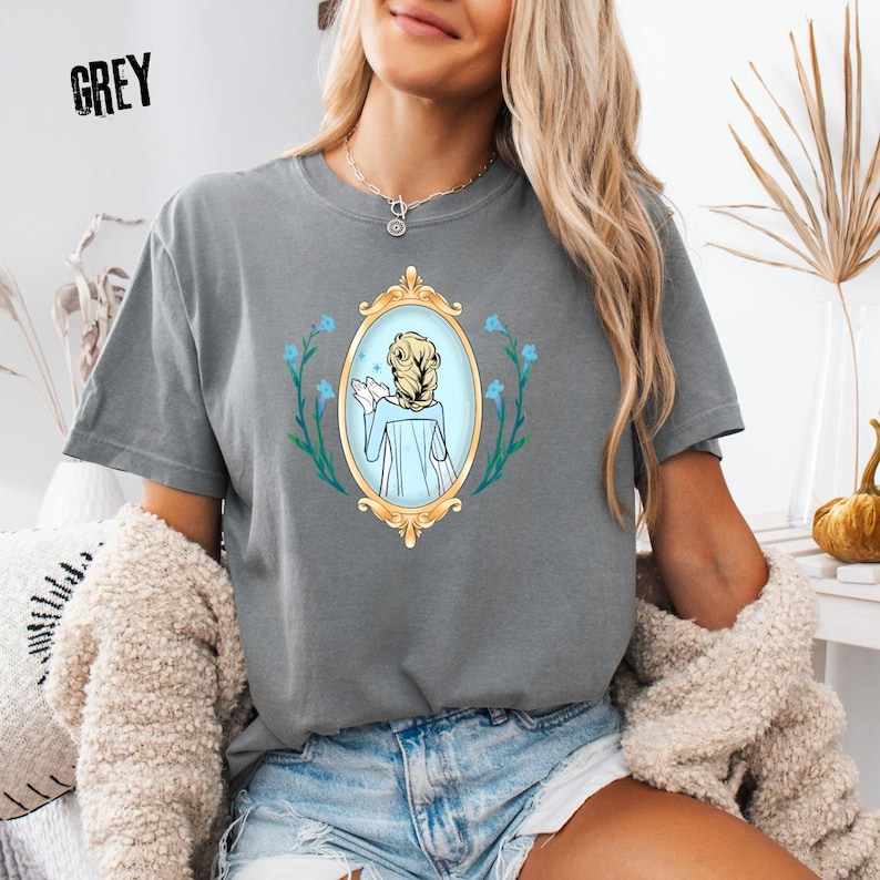 May include: Grey t-shirt featuring a cartoon illustration of a woman in a mirror, surrounded by blue flowers. The mirror has a gold frame. The word "GREY" is printed in the upper left corner.