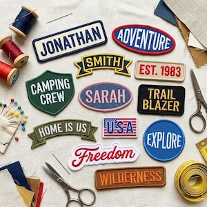 May include: A collection of embroidered fabric patches featuring various shapes and text including JONATHAN, ADVENTURE, SMITH, EST. 1983, CAMPING CREW, SARAH, TRAIL BLAZER, HOME IS US, USA, EXPLORE, Freedom, and WILDERNESS, arranged with sewing supplies like thread.