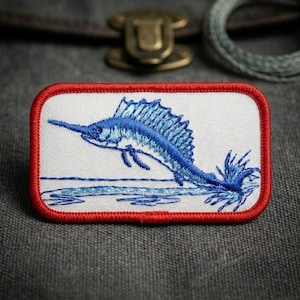 May include: A rectangular embroidered patch featuring a blue sailfish leaping above stylized blue water waves. The patch has a white background and is framed by a thick, vibrant red stitched border, creating a distinct contrast in color.