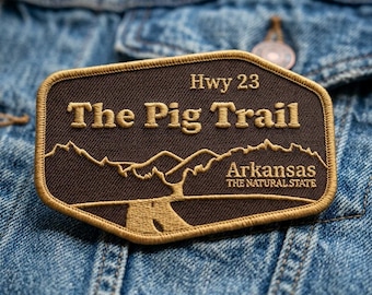 May include: A rectangular embroidered patch with rounded corners featuring a dark brown background and gold border. It displays the text Hwy 23, The Pig Trail, and Arkansas The Natural State, along with a gold stitched mountain and road graphic.