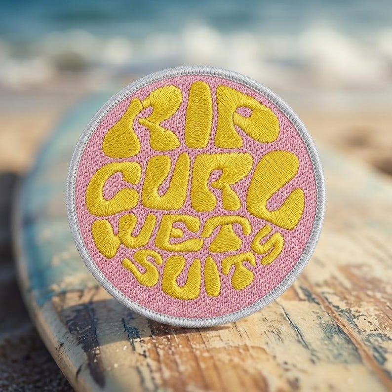 May include: A circular embroidered patch with a pink background and a white stitched border. The text RIP CURL WET SUITS is embroidered in a bold, yellow, rounded font across the center of the patch, filling the circular space completely.