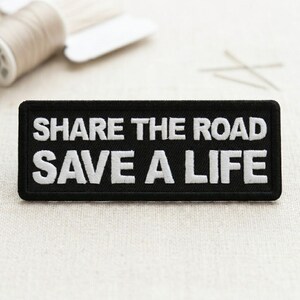 May include: Black rectangular patch with white embroidered text that reads "SHARE THE ROAD SAVE A LIFE". The patch is likely for a jacket or bag. Sewing supplies are in the background.