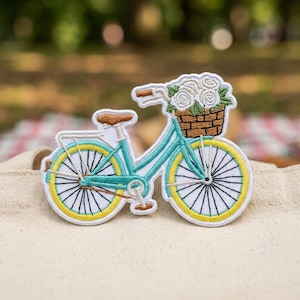 May include: Embroidered patch of a teal bicycle with yellow-trimmed wheels and a brown basket filled with white roses. The bicycle has a brown seat and handlebars. The patch is on a beige surface.