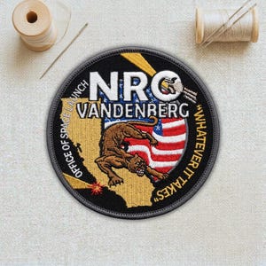 May include: A round embroidered patch with the text "NRO VANDENBERG" and a cougar over an American flag. The patch also features the text "OFFICE OF SPACE LAUNCH" and "WHATEVER IT TAKES" around the border. The patch is black, brown, yellow, white, and red.
