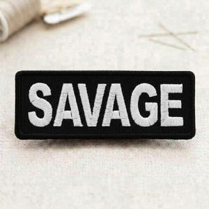 May include: Black rectangular patch with the word "SAVAGE" embroidered in white. The patch has a black border and is displayed on a neutral-colored surface. The bold text is the focal point.