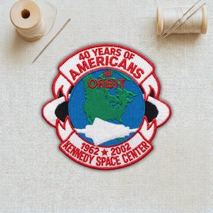 May include: Embroidered patch commemorating "40 Years of Americans in Orbit" from 1962-2002, featuring a globe with North America in green, a blue background, and the text "Kennedy Space Center". Includes spools of thread and a needle.