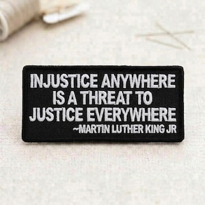 May include: A black rectangular patch with white embroidered text. The text reads "INJUSTICE ANYWHERE IS A THREAT TO JUSTICE EVERYWHERE" and "~MARTIN LUTHER KING JR". The patch is likely for clothing or accessories.