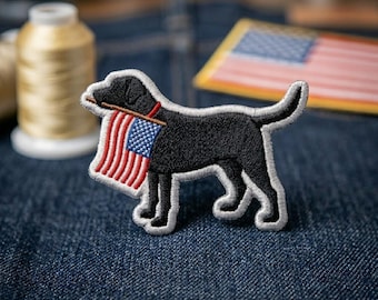 May include: An embroidered patch featuring a black Labrador retriever in profile, facing left. The dog holds a small American flag in its mouth. The patch has a white stitched border and is rendered in black, red, white, and blue thread colors.
