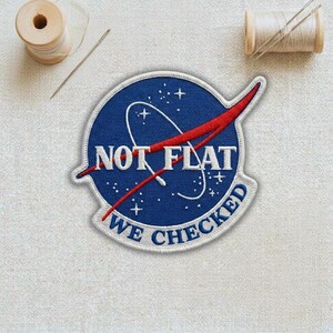 May include: Embroidered patch with a blue background, white stars, and a red swoosh design. The patch reads "NOT FLAT WE CHECKED" in white lettering. Two spools of thread and a needle are in the background.
