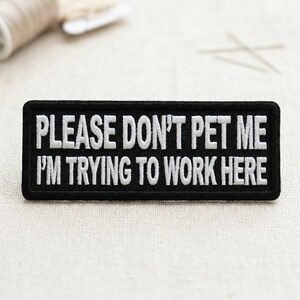 May include: A rectangular black patch with white embroidered text that reads "PLEASE DON'T PET ME I'M TRYING TO WORK HERE". The patch has a black border and is likely designed to be sewn or ironed onto clothing or accessories.