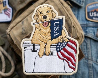 May include: An embroidered patch featuring a golden retriever dog resting on a white cooler. The dog holds a small blue flag with the text USA in its mouth. A red, white, and blue American flag is draped over the side of the cooler. The patch has a cream-colored border and detailed stitching throughout the design.