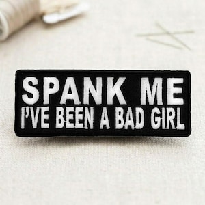 May include: Black rectangular patch with white embroidered text that reads "SPANK ME I'VE BEEN A BAD GIRL". The patch is likely for clothing or accessories.