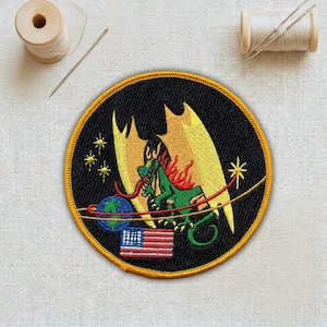 May include: Embroidered patch featuring a green dragon with yellow wings and red flames, set against a black background. The design includes a globe, the American flag, and stars, with a gold border. The patch is round.