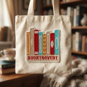 May include: Beige canvas tote bag featuring an embroidered patch of colorful books. The patch includes books in shades of red, orange, teal, and brown. The word "BOOKTROVERT" is embroidered in red below the books. The tote bag has matching beige handles.