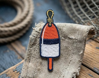 May include: Embroidered patch featuring a nautical buoy design. The patch has a red and white color scheme with a navy blue stripe. The buoy is topped with a yellow loop and a navy blue outline. The patch is on a tan fabric.