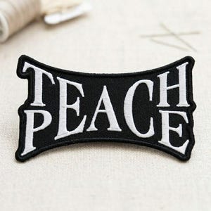 May include: A black embroidered patch with the word "TEACH PEACE" in white, stylized lettering. The patch has a curved rectangular shape with a black border. The background is a neutral, light-colored fabric.