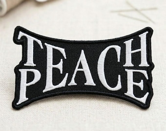 May include: A black embroidered patch with the word "TEACH PEACE" in white, stylized lettering. The patch has a curved rectangular shape with a black border. The background is a neutral, light-colored fabric.