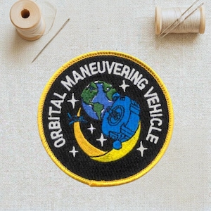 May include: Embroidered patch with the text "ORBITAL MANEUVERING VEHICLE". The patch features a blue and green Earth, a blue spacecraft, and a yellow crescent moon with stars on a black background, with a yellow border.