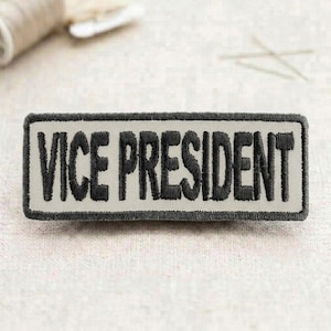 May include: Embroidered patch with the words "VICE PRESIDENT" in bold, black letters. The patch has a rectangular shape with a light gray background and a dark gray border. The patch is likely for a uniform or clothing.