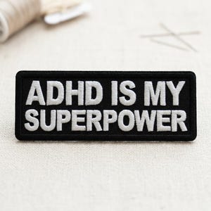 May include: Black rectangular patch with white embroidered text that reads "ADHD IS MY SUPERPOWER." The patch has a black border and is likely made of fabric. The background is a neutral color, and sewing supplies are visible.