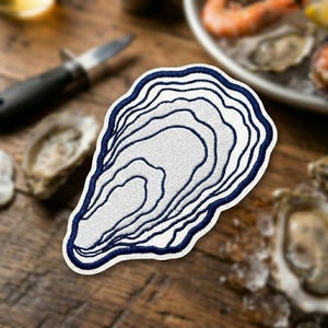 May include: Embroidered patch in the shape of an oyster, featuring a white interior with navy blue contour lines and a navy blue border. The patch is on a wooden surface, with oyster shells and a knife in the background.