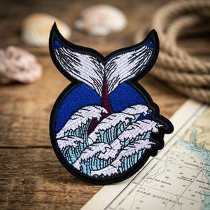 May include: A circular embroidered patch featuring a white whale tail extending upward from a deep blue background. Below the tail, stylized white and light blue ocean waves are depicted with black outlines, all bordered by a thick black edge.