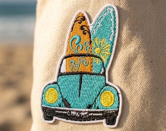 May include: An embroidered patch featuring a teal car with yellow headlights and a black bumper. Two surfboards are strapped to the roof: one orange with black swirls and one teal with a yellow sun design. The patch is attached to beige fabric.