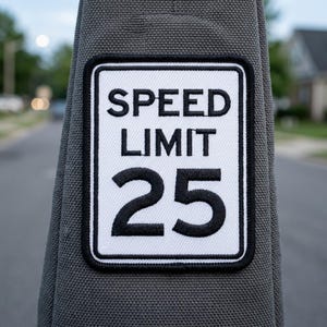 May include: A rectangular patch with a white background and black border, featuring the text "SPEED LIMIT 25" in bold black letters. The patch is sewn onto a gray fabric.