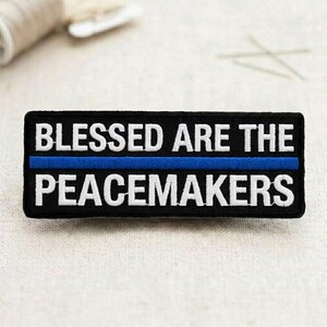 May include: A rectangular black patch with white embroidered text that reads "BLESSED ARE THE PEACEMAKERS." A blue line is embroidered across the center of the patch. The patch is likely an accessory.