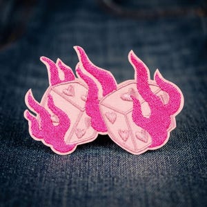 May include: An embroidered patch featuring two light pink dice with heart-shaped pips, each engulfed in vibrant hot pink flames. The design is rendered with detailed stitching, showcasing a clear contrast between the soft pink dice and the bright pink fire.
