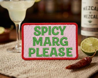 May include: A margarita cocktail with a lime wedge, a red chili pepper, and a patch that reads "Spicy Marg Please" in green letters on a pink background. A bottle of mezcal is in the background.