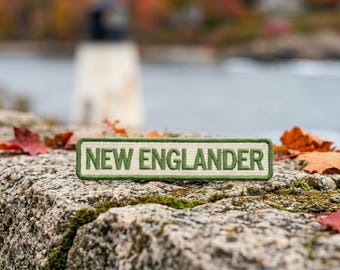 May include: A rectangular patch with the text "NEW ENGLANDER" in green, embroidered on a cream-colored background. The patch is set against a blurred background of a lighthouse, water, and fall foliage.