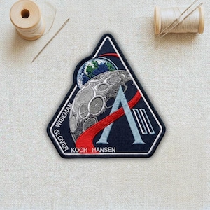 May include: A navy blue embroidered patch with a white border, featuring a stylized Earth and moon design. The patch includes the names GLOVER, WISEMAN, KOCH, and HANSEN. A red and light blue design is also present.