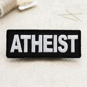 May include: A black rectangular patch with the word "ATHEIST" embroidered in white, bold capital letters. The patch is likely made of fabric and designed to be sewn or ironed onto clothing or accessories.