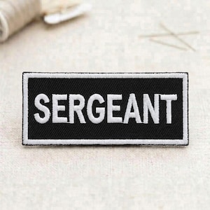 May include: A black rectangular patch with the word "SERGEANT" embroidered in white block letters. The patch has a white border and is likely for uniform identification.