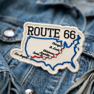May include: An embroidered patch shaped like the United States map with a cream background and blue border. It features the black text ROUTE 66, a red stitched path, and white stars marking cities including Chicago, St. Louis, Oklahoma, Amarillo, Santa Fe, and Los Angeles.
