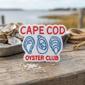 May include: A white embroidered patch with a scalloped edge featuring the red text CAPE COD at the top and OYSTER CLUB at the bottom. Between the text are three blue embroidered outlines of oyster shells arranged horizontally on the fabric.