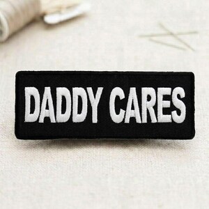 May include: Black rectangular patch with the embroidered text "DADDY CARES" in white. The patch is likely designed to be sewn or ironed onto clothing or accessories. The design is simple and bold.
