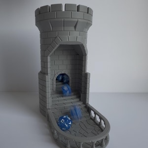 May include: A gray, 3D-printed dice tower in the shape of a castle. Blue dice are rolling down the stairs and onto the balcony. The tower has a brick pattern and a crenelated top. The balcony has a stone pattern.