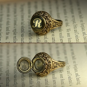 Può includere: Un anello dorato con un coperchio a cerniera, decorato con un elaborato motivo floreale e la lettera "R" incisa sulla parte superiore. L'anello è aperto, rivelando un piccolo scomparto all'interno.