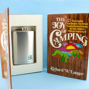 May include: A book titled "The Joy of Camping: The Complete Four-Seasons, Five-Senses Practical Guide to Enjoying the Great Outdoors (Without Destroying It)" by Richard W. Langer. The book is open to reveal a silver flask with a green "R" engraved on it.