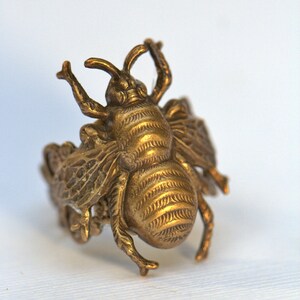 May include: A gold-toned bee ring with intricate details. The bee is depicted with wings, legs, and a segmented body. The ring is made of metal and has a textured finish.