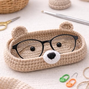 Peut inclure: Un porte-lunettes en forme d'ours crocheté de couleur beige clair, avec des lunettes à monture noire à l'intérieur. Le support a deux petites oreilles et un nez blanc et noir. D'autres fournitures de bricolage sont visibles.