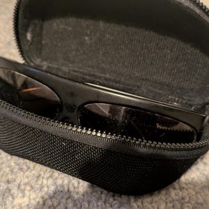 May include: Black sunglasses with dark lenses rest inside an open, black zippered case. The case is made of a textured fabric. The sunglasses have a thick frame. The case is on a light gray surface.