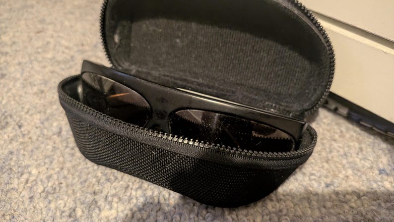 May include: Black sunglasses with dark lenses rest inside an open, black zippered case. The case is made of a textured fabric. The sunglasses have a thick frame. The case is on a light gray surface.