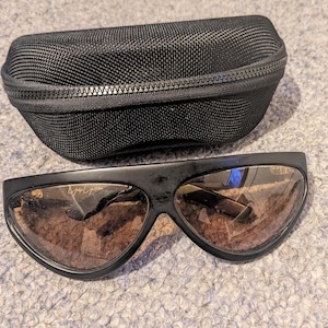 May include: Black sunglasses with brown lenses and a matching black zippered case. The sunglasses have a unique, curved design and a small logo on the frame. The case is made of a textured, woven material.