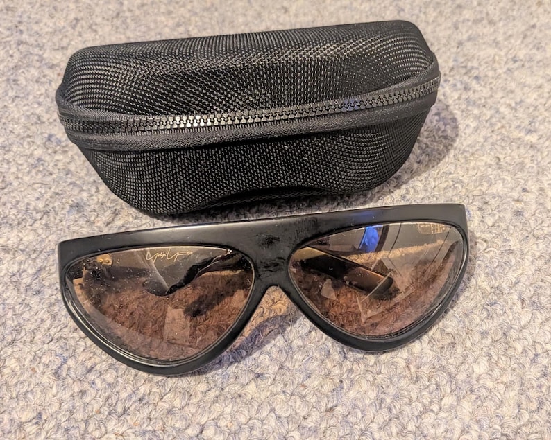 May include: Black sunglasses with brown lenses and a matching black zippered case. The sunglasses have a unique, curved design and a small logo on the frame. The case is made of a textured, woven material.