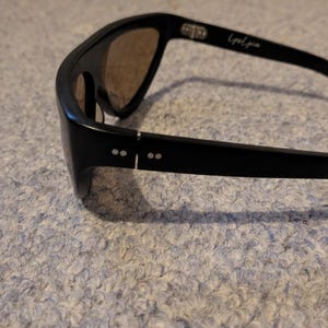 May include: Black sunglasses with brown lenses. The sunglasses have a sleek, modern design with a thick frame and curved temples. The text "Liquid Zone" is printed on the temple. The sunglasses are resting on a textured surface.