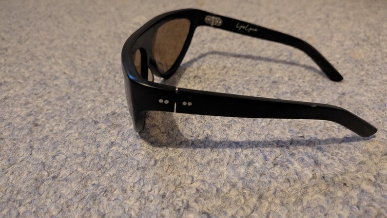 May include: Black sunglasses with brown lenses. The sunglasses have a sleek, modern design with a thick frame and curved temples. The text "Liquid Zone" is printed on the temple. The sunglasses are resting on a textured surface.