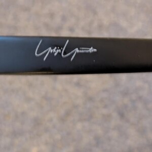 May include: Close-up of a black eyeglass arm with a silver hinge and the designer's signature in silver. The arm is rectangular and has a matte finish.